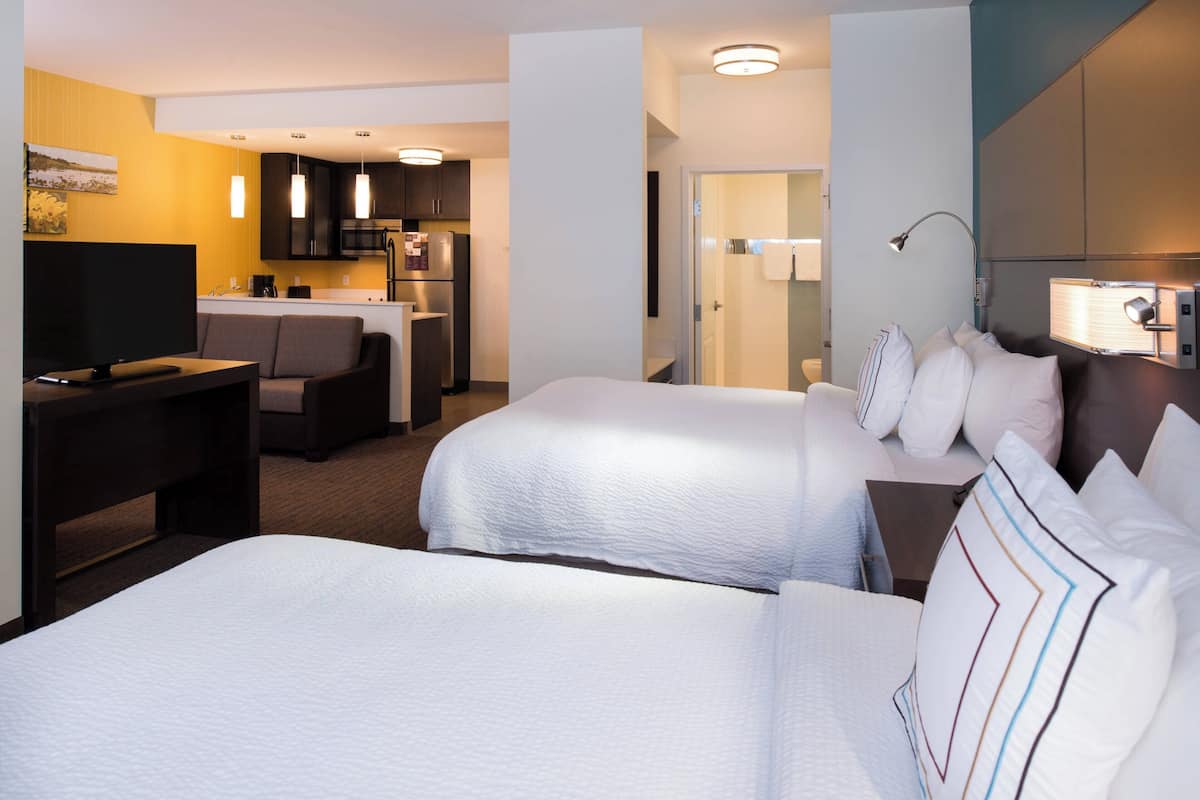 A hotel room with two beds, a desk, a TV, and a kitchenette.