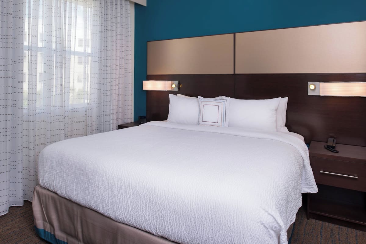 A large bed with a white comforter, a wooden headboard, two bedside tables with lamps, and a window with sheer curtains.