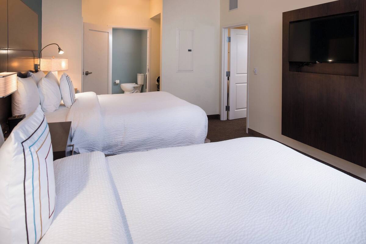 A hotel room with two beds, a TV, a desk with a lamp, and a bathroom visible through an open door.