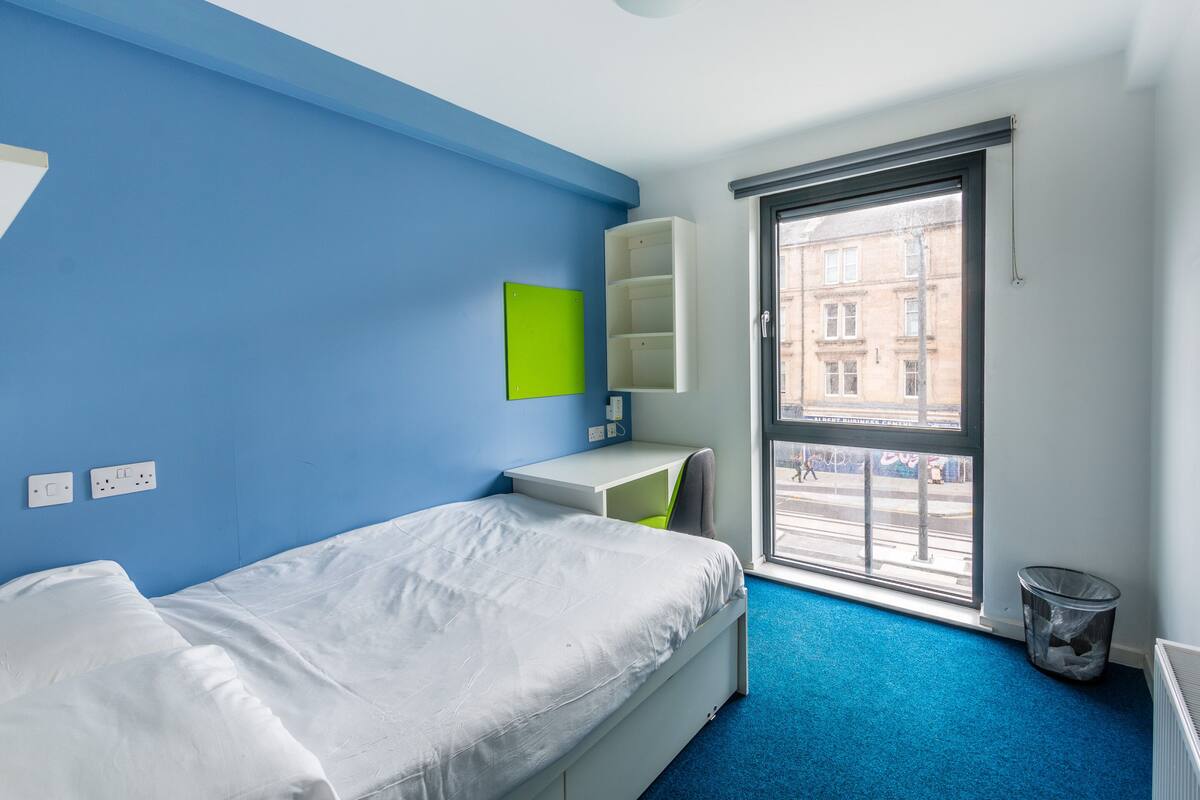 A bedroom with a bed, a desk, a chair, a window, and a radiator.