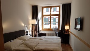 Une chambre d’hôtel avec un grand lit, un bureau, une chaise, une lampe, un ventilateur et une fenêtre avec des rideaux.