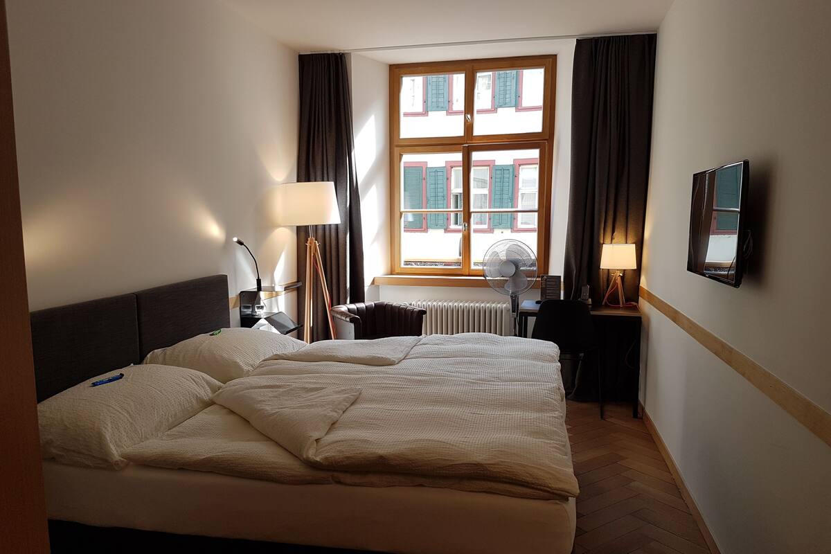Une chambre d’hôtel avec un grand lit, un bureau, une chaise, une lampe, un ventilateur et une fenêtre avec des rideaux.