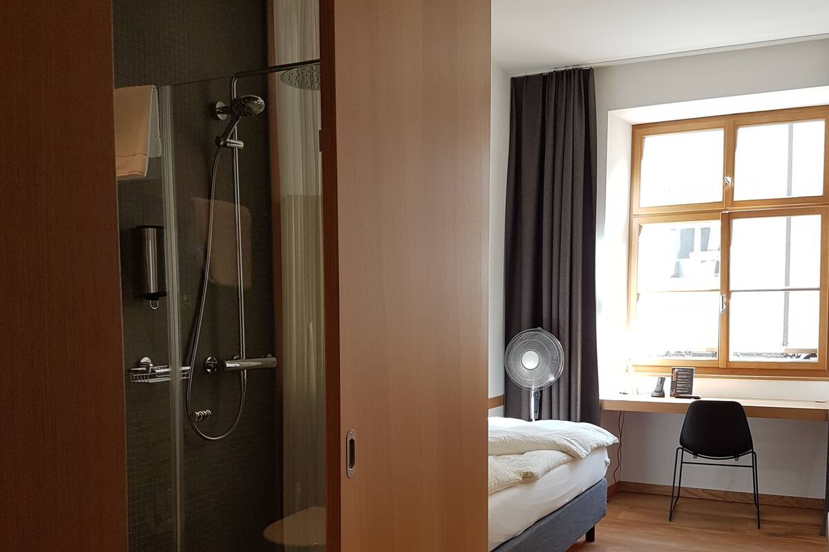 Une chambre moderne dotée d’une douche en verre, d’un lit recouvert d’une couette blanche, d’un bureau avec une chaise et d’une fenêtre avec des rideaux.