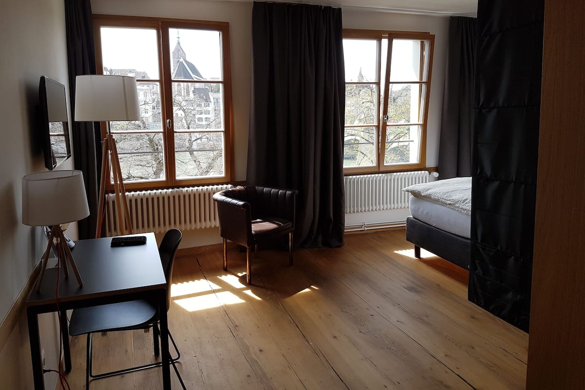 Une pièce avec un plancher en bois, un bureau avec une lampe, une chaise, un lit et de grandes fenêtres avec des rideaux.