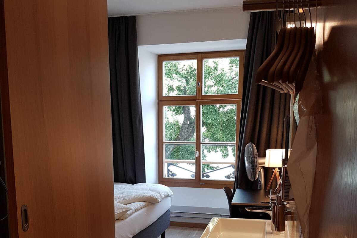 Une chambre d’hôtel avec un lit, une fenêtre avec des rideaux, un lavabo et un ventilateur.