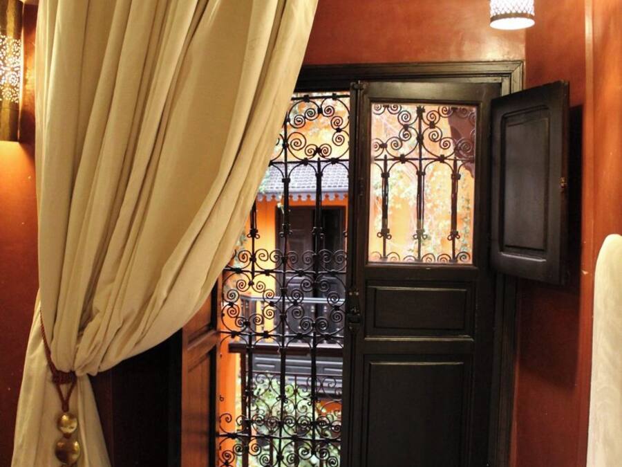 Una habitación con una cama, una ventana con cortina, un aplique en la pared y una puerta con reja de metal.