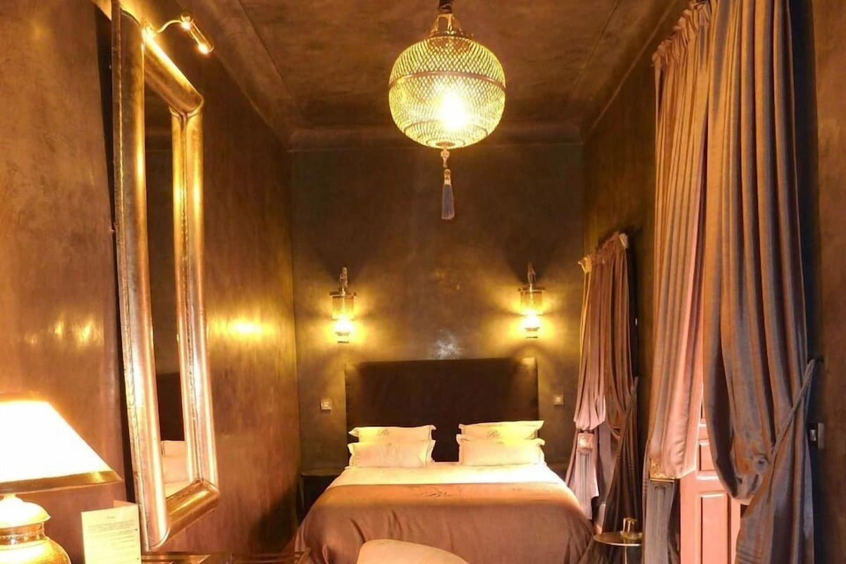 Une chambre d’hôtel avec un lit, une chaise, un bureau avec une lampe, un miroir et un lustre.