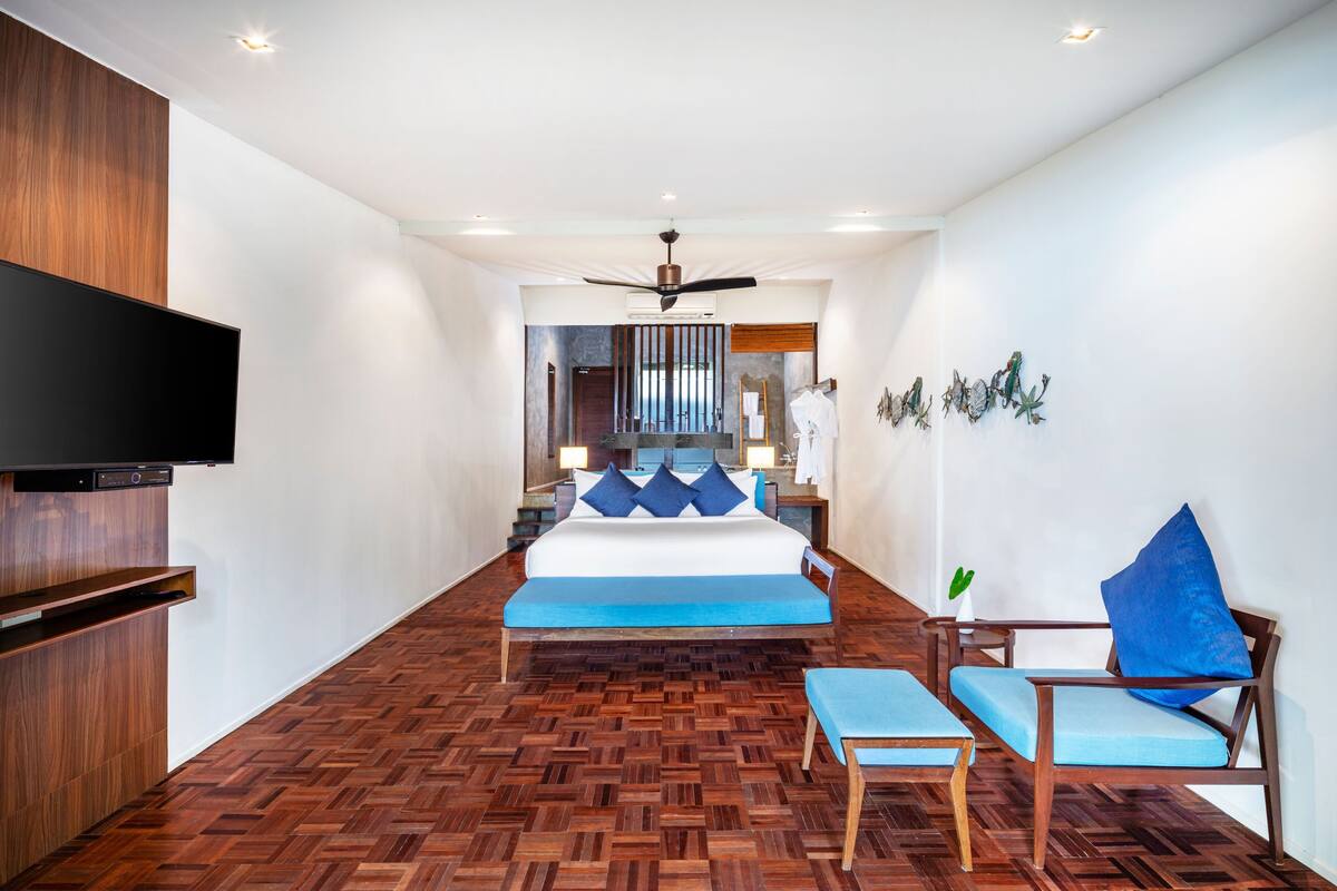 A modern hotel room with a wooden floor, a bed with blue bedding, a TV, a chair, and a ceiling fan.
