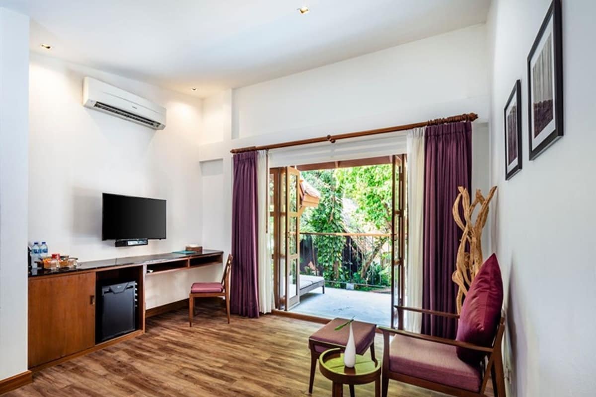 A room with a wooden floor, a desk with a TV, a chair, a sofa, and a view of greenery outside.