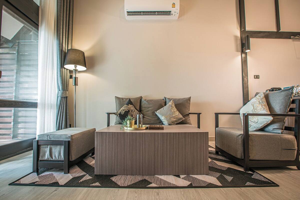 A modern living room with a sofa, a coffee table, and a chair. There is a lamp, a plant, and a small table with a vase. An air conditioning unit is mounted on the wall.