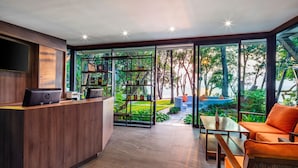 A modern reception area with a wooden desk, a HP laptop, and a bookshelf. There is a large glass door leading to an outdoor garden area with a blue seating area. - The Monttra Pattaya (Pattaya)