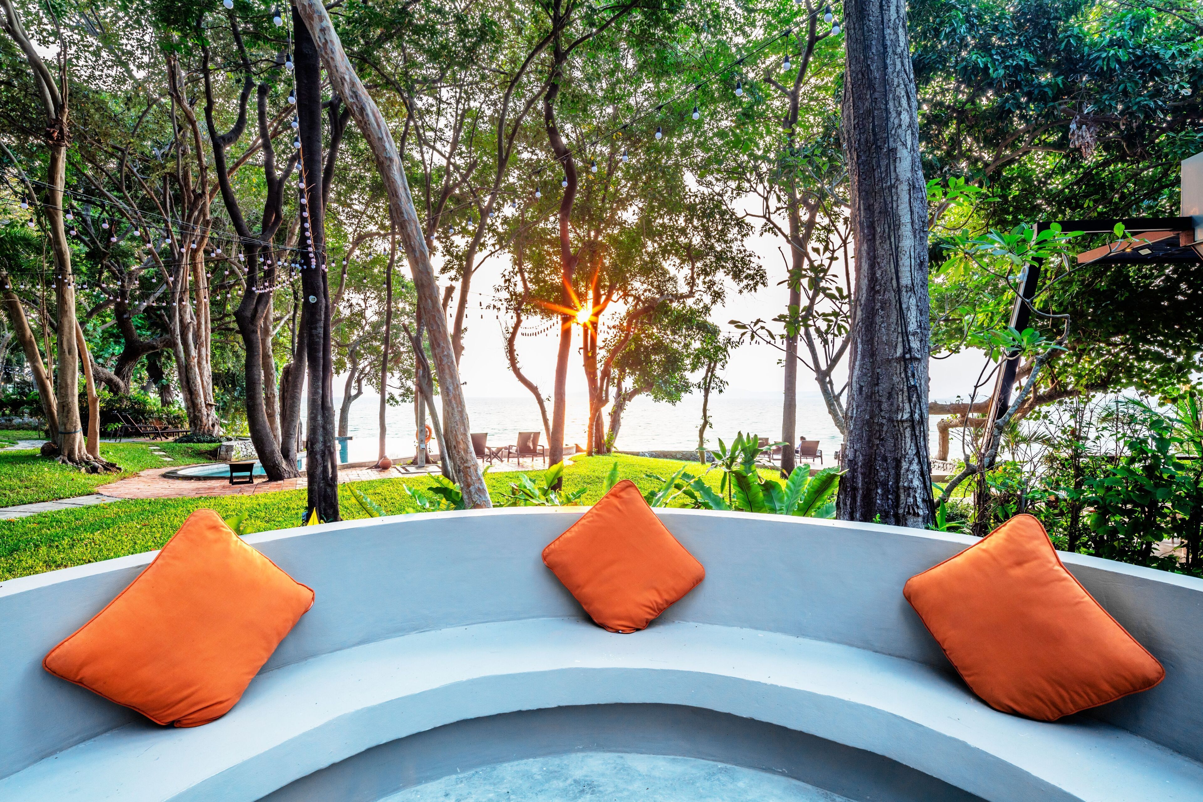 Un banc incurvé avec des coussins orange, entouré d’une végétation luxuriante et d’arbres, et offrant une vue sur une étendue d’eau.