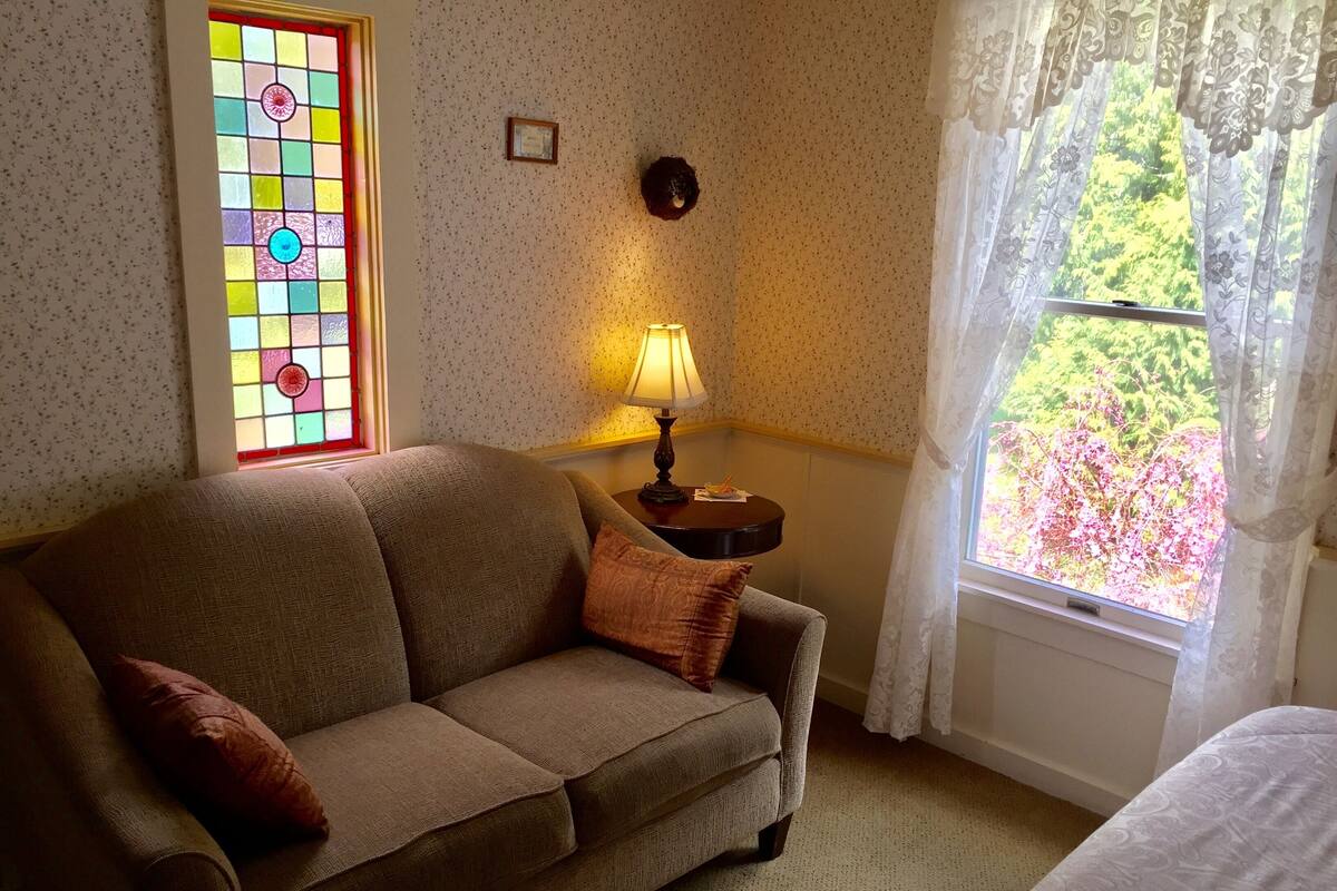 Ein gemütliches Zimmer mit einem braunen Sofa, einem kleinen Tisch mit Lampe, einem Buntglasfenster und Blick auf Grün durch ein Fenster mit durchsichtigen Vorhängen.