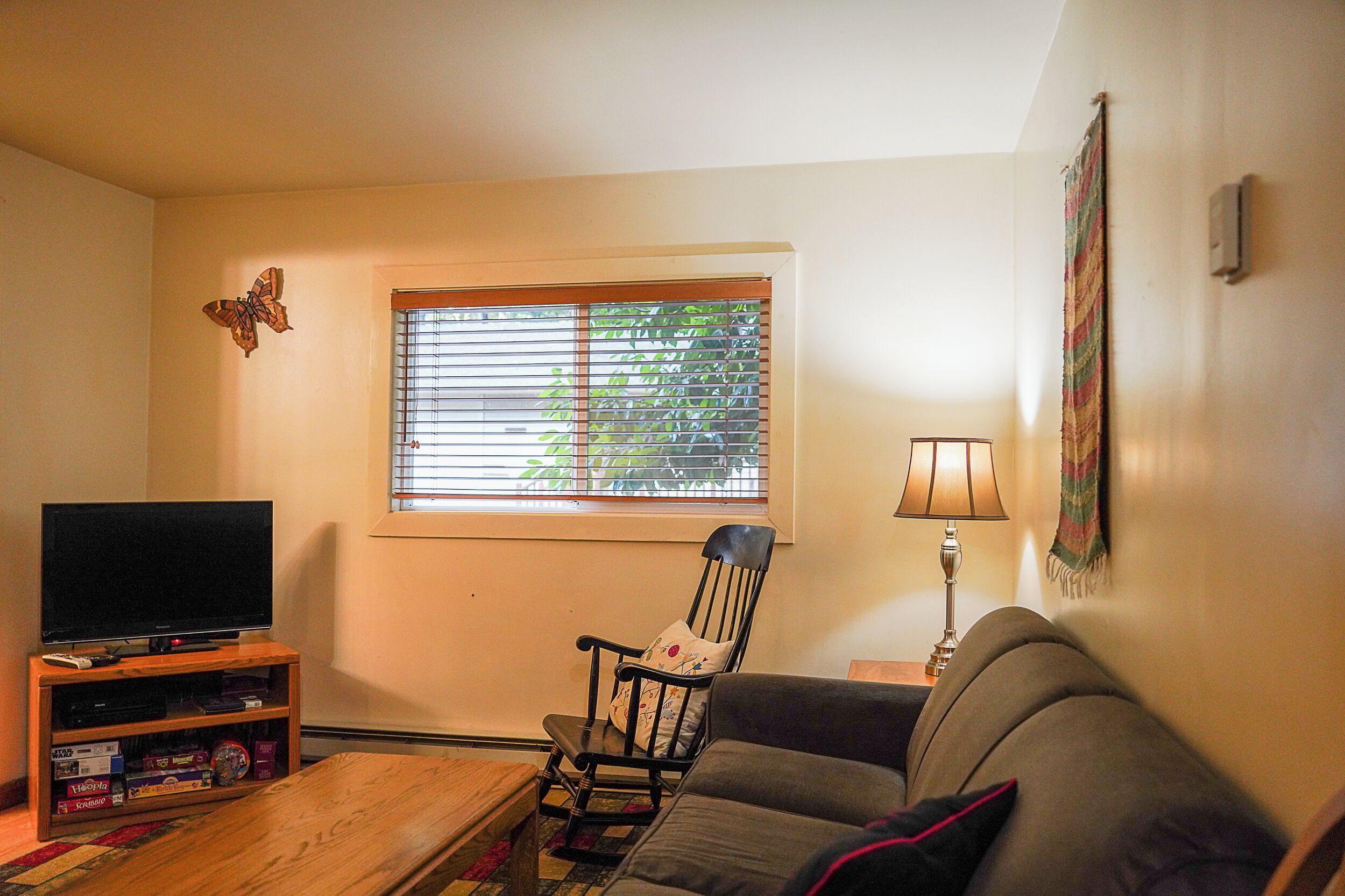 A living room with a television on a wooden stand, a rocking chair, a sofa, and a window with blinds.