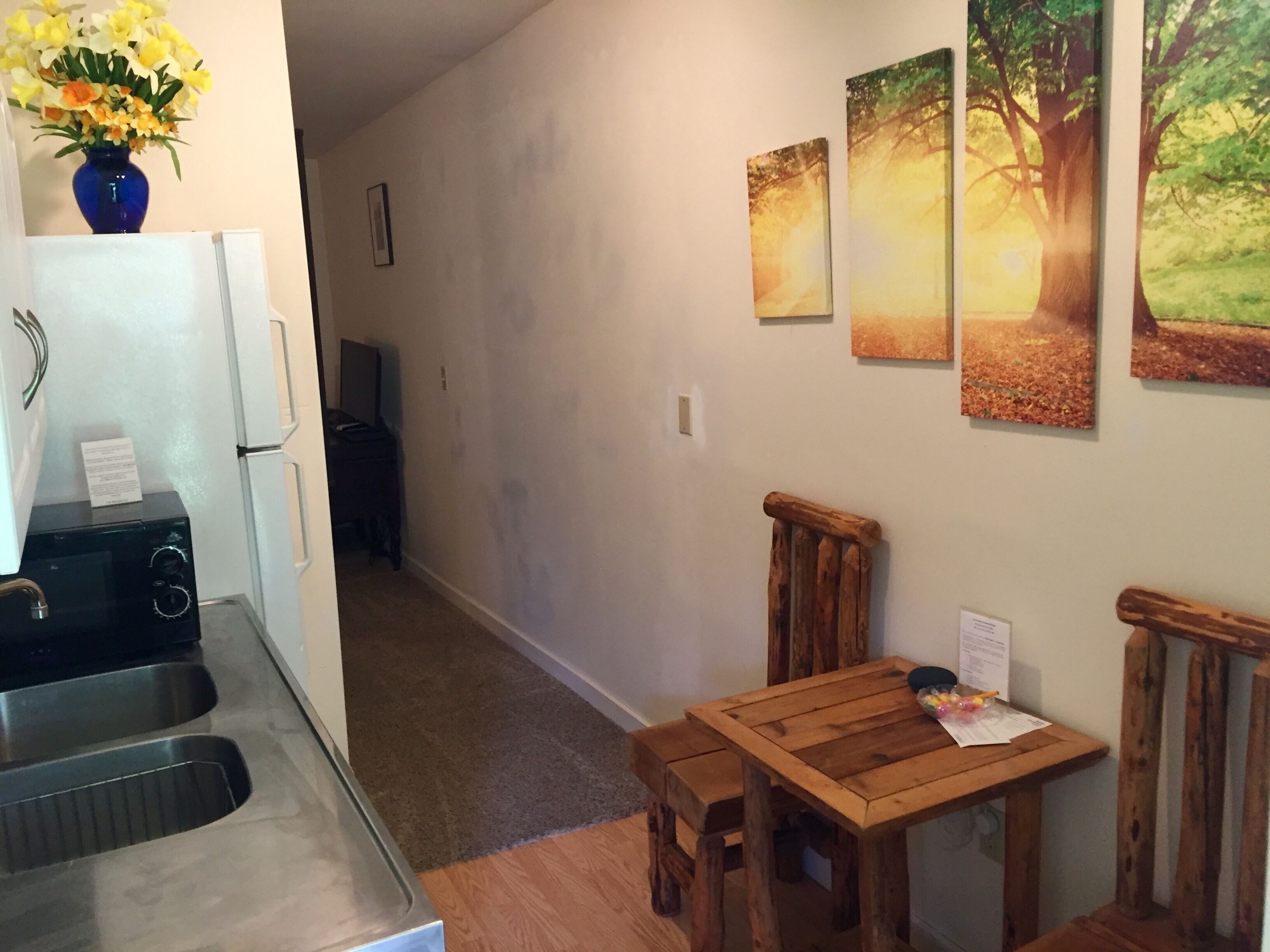 A kitchen with a microwave, sink, and a vase of flowers. There is a wooden table with chairs and a painting of a forest scene on the wall.