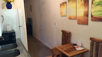 A kitchen with a microwave, sink, and a vase of flowers. There is a wooden table with chairs and a painting of a forest scene on the wall.