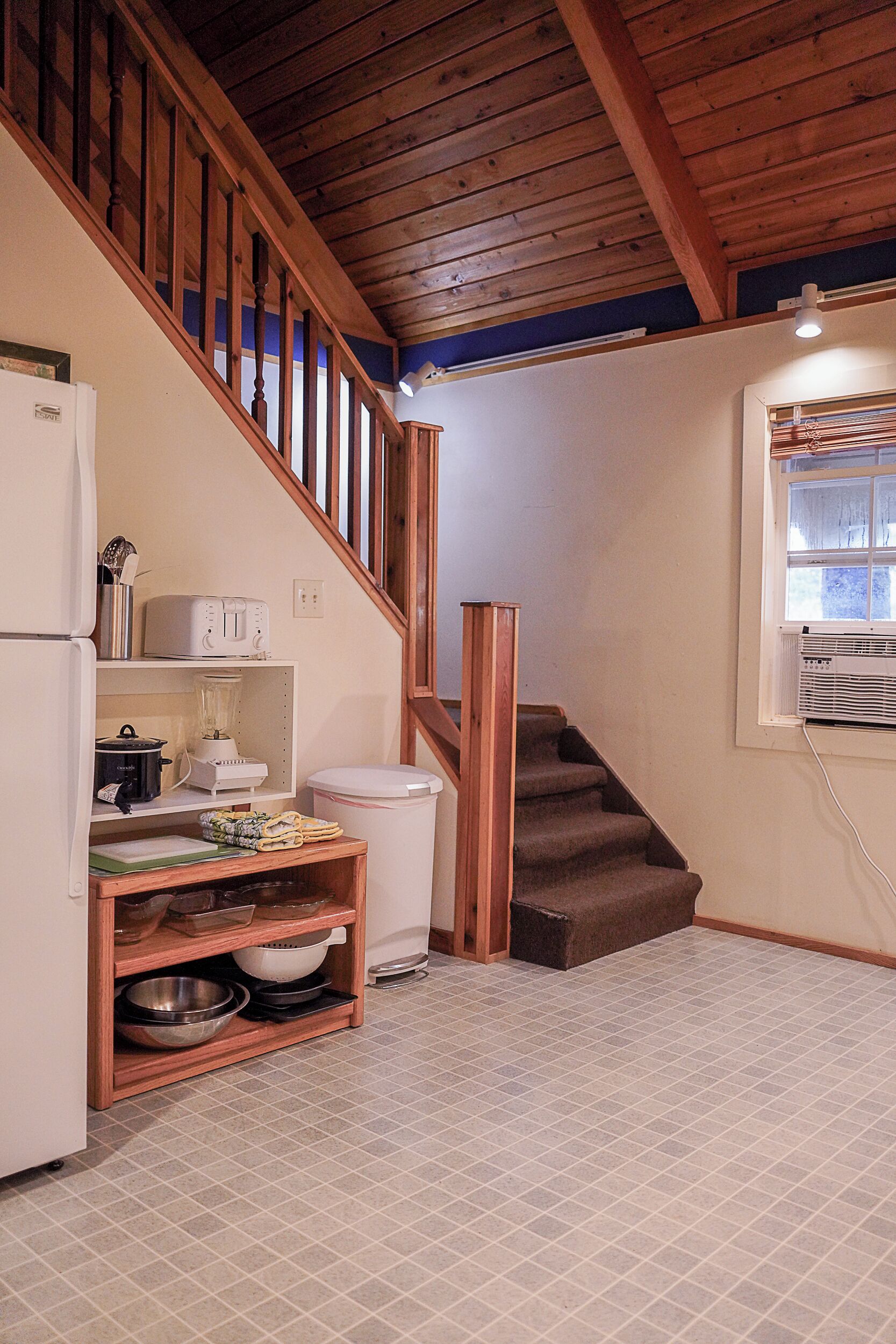 A kitchen with a wooden staircase, a refrigerator, a toaster, a blender, and a trash bin.