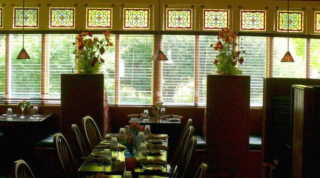 A dining area with a long table set for a meal, large windows with stained glass, and potted plants.