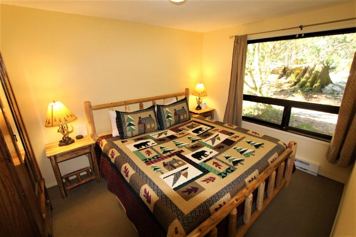 Ein gemütliches Zimmer mit einem hölzernen Bett, zwei Nachttischlampen, einem Fenster mit Blick auf Bäume und einem Bettbezug mit Tiermotiven.