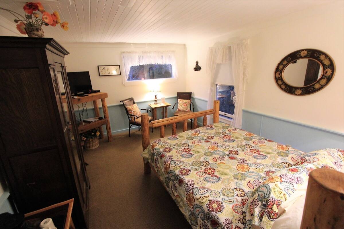 Ein gemütliches Schlafzimmer mit einem Bett, einer Holzkommode, einem Schreibtisch mit Stuhl, einem Fenster mit Vorhängen und einem Spiegel an der Wand.