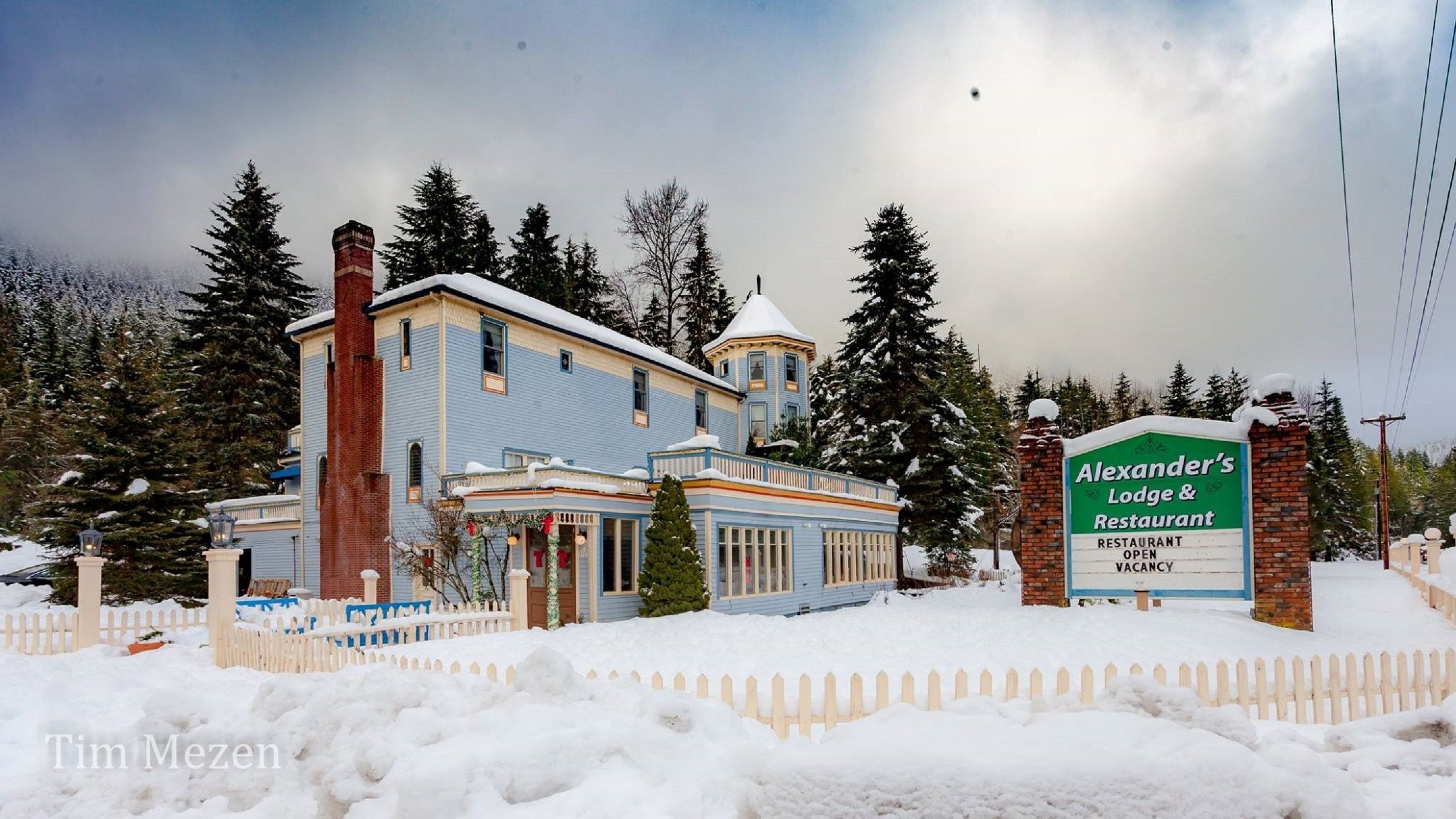 Een gebouw met twee verdiepingen, een opvallende schoorsteen, een kleine toren en een bordje met Alexander's Lodge & Restaurant erop.
