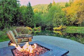 Un brasero con leña ardiendo, rodeado de grava, un banco de madera y vistas a un lago y un bosque.