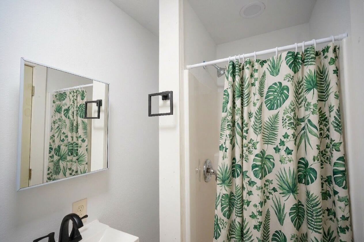 Une salle de bain avec un miroir, un lavabo et un rideau de douche orné d’un motif de feuilles tropicales.