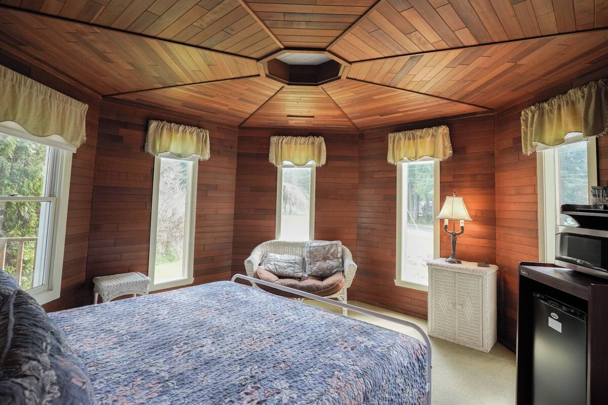 Ein Zimmer mit Holzwänden, einem Bett mit Blumenmuster, einem Sessel, einer Lampe und einem Kühlschrank.