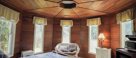 A room with wooden walls, a bed with a floral bedspread, a chair, a lamp, and a refrigerator.