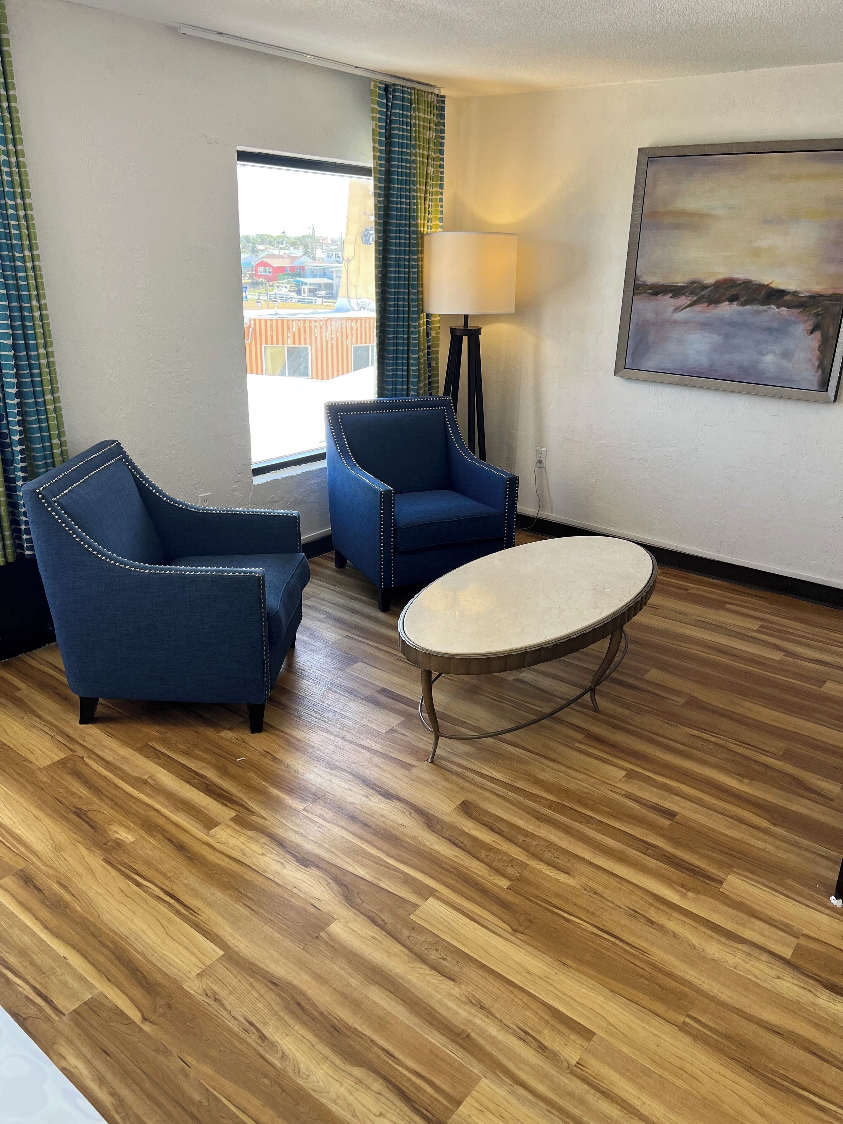 Une chambre d’hôtel avec deux fauteuils bleus, une table ronde, un lampadaire et un tableau accroché au mur.