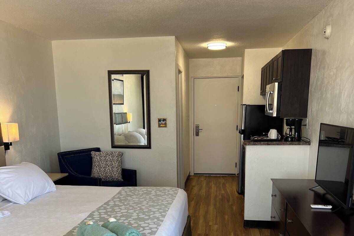 Ein Hotelzimmer mit einem Bett, einem Schreibtisch, einem Stuhl, einem kleinen Tisch, einem Spiegel und einer Küchenzeile.