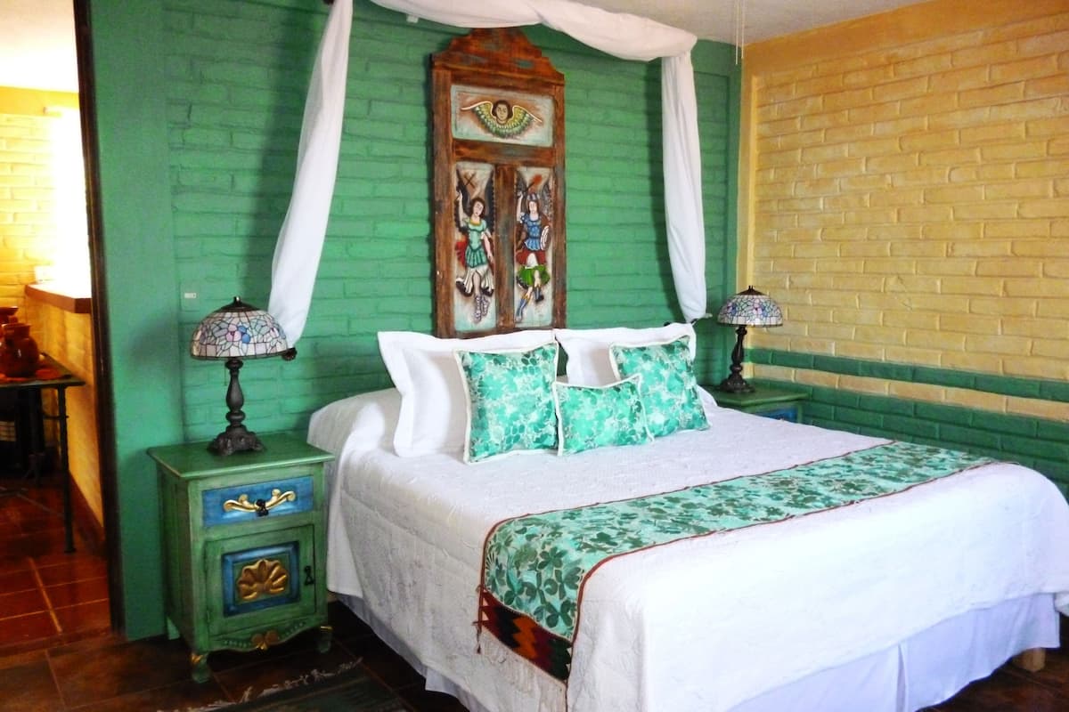 A room with a bed, green walls, a wooden headboard, a nightstand with a lamp, and a patterned bedspread.
