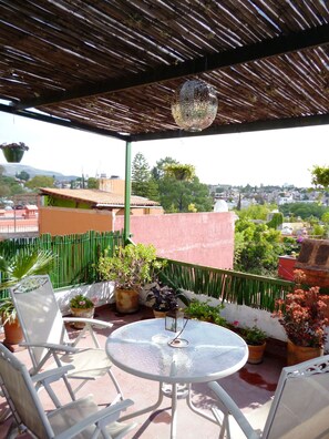 A covered outdoor seating area with a table and chairs, surrounded by potted plants and a view of the cityscape.