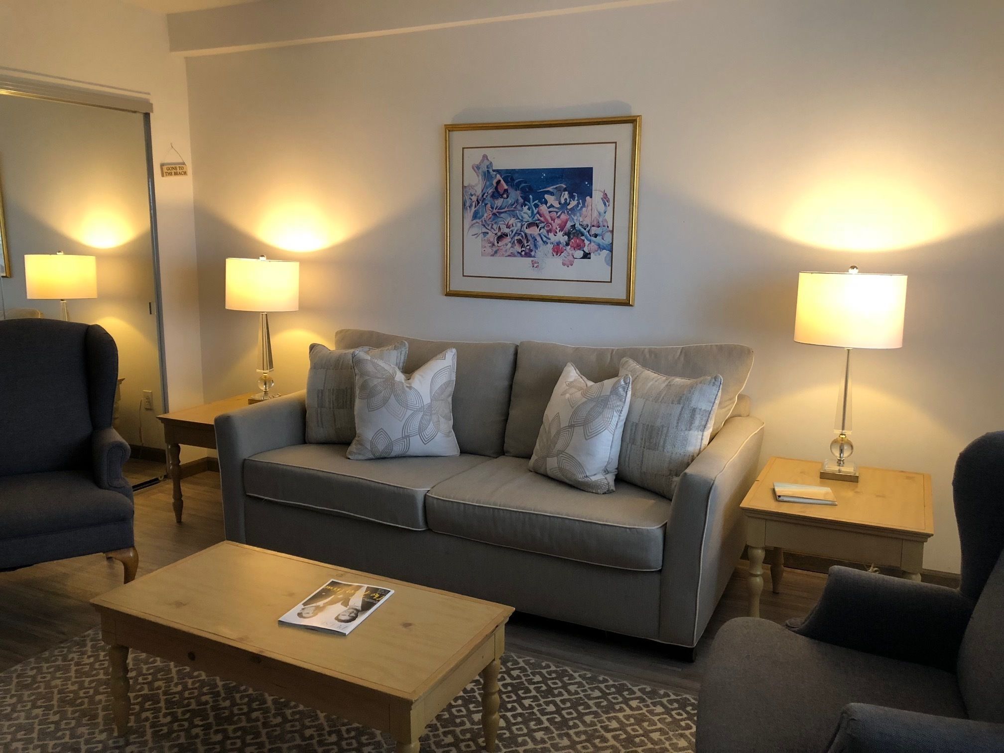 Un soggiorno con un divano grigio, due poltrone, un tavolino da caffè e due comodini con lampade. Al di sopra del divano, c'è un quadro incorniciato appeso al muro.