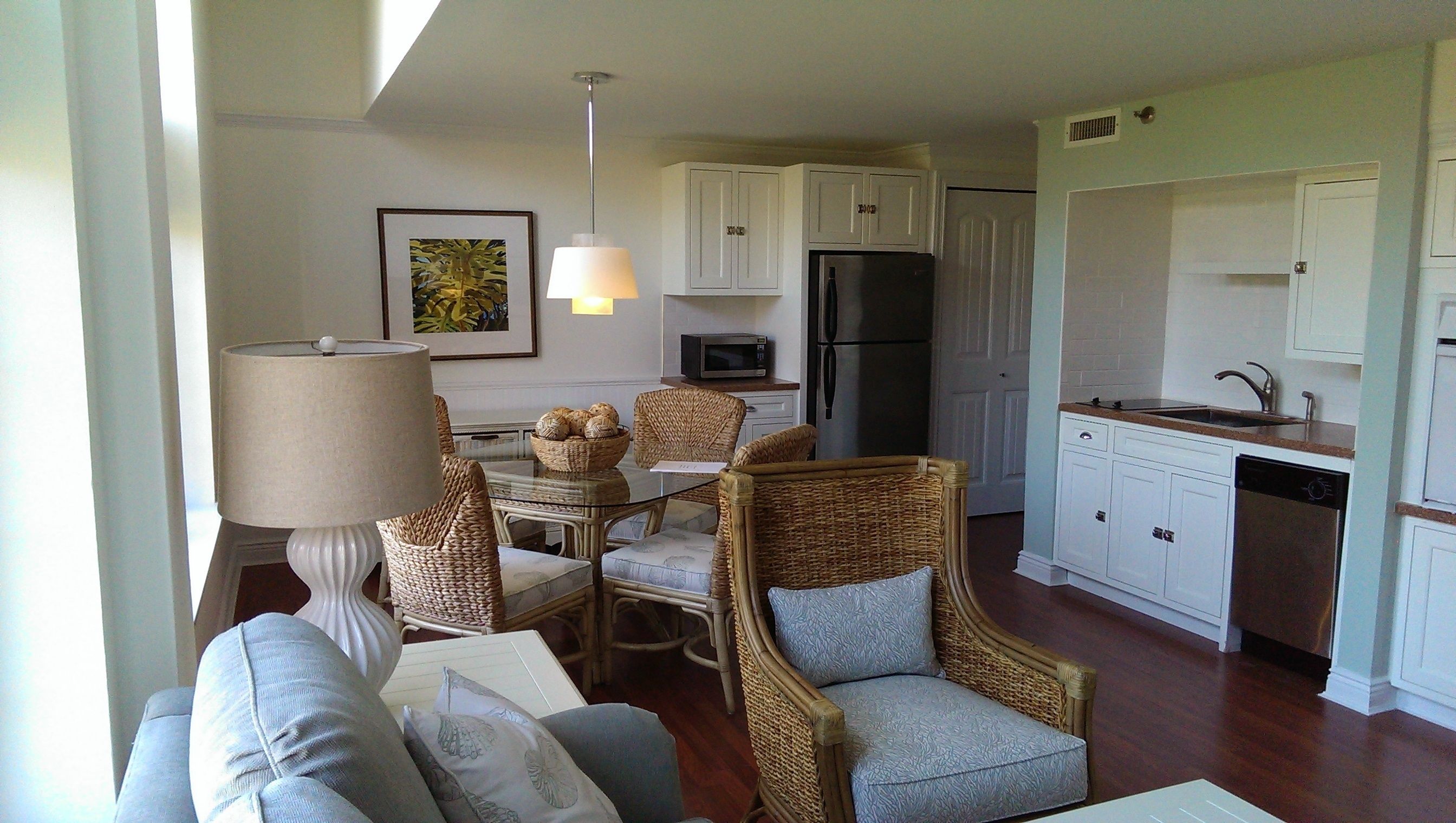 A living room with a sofa, chairs, a table, a lamp, and a dining area with wicker chairs and a glass table. The kitchen has white cabinets, a refrigerator, a microwave, and a dishwasher.