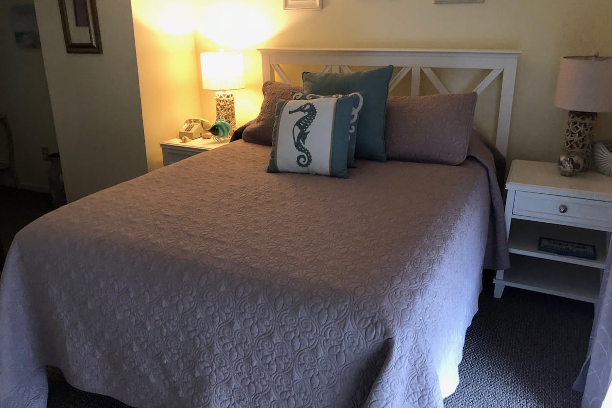 Una cama bien hecha con una colcha estampada, un cojín decorativo y una lámpara de noche.
