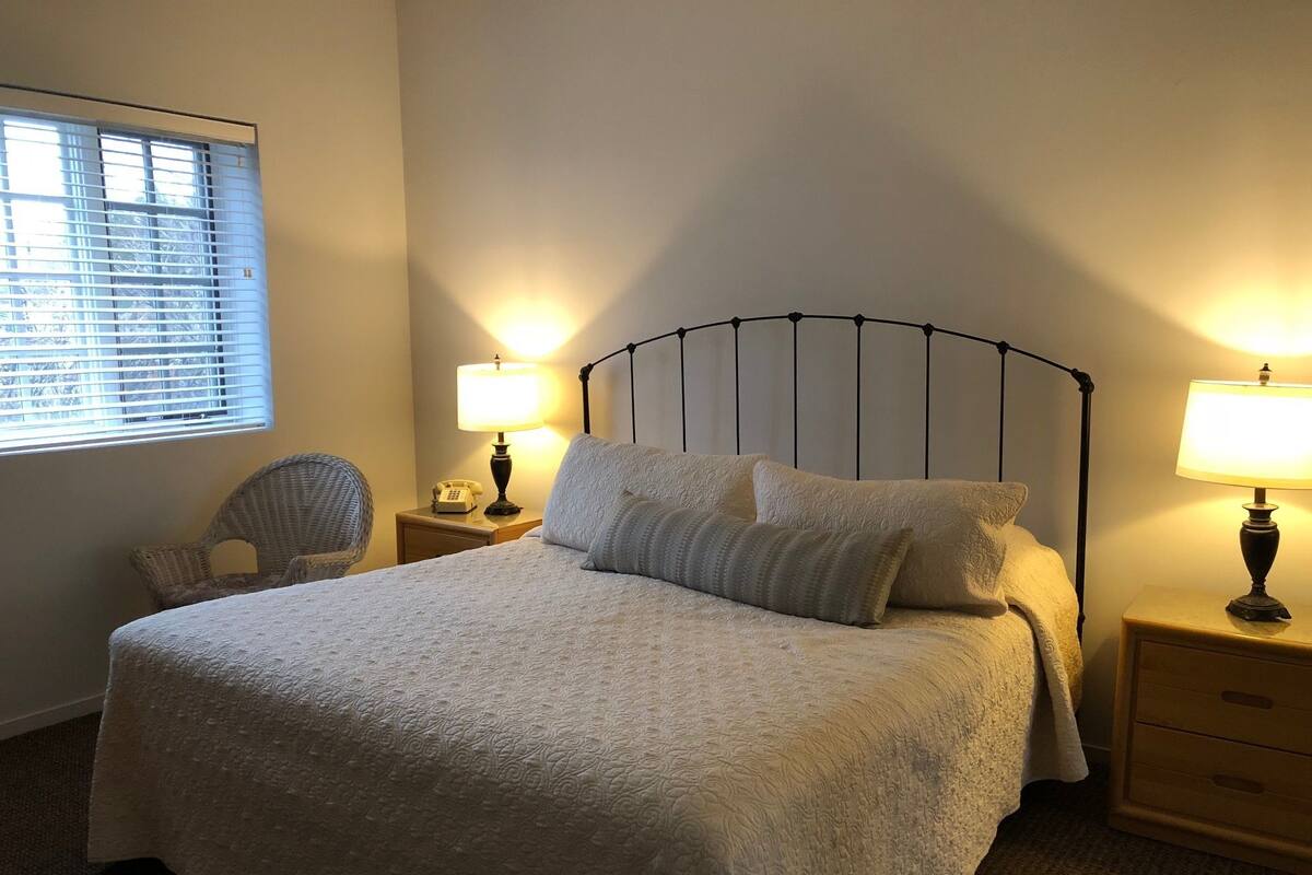 Una cama bien hecha con cabecera, dos mesitas de noche con lámparas, una silla y una ventana con persianas.