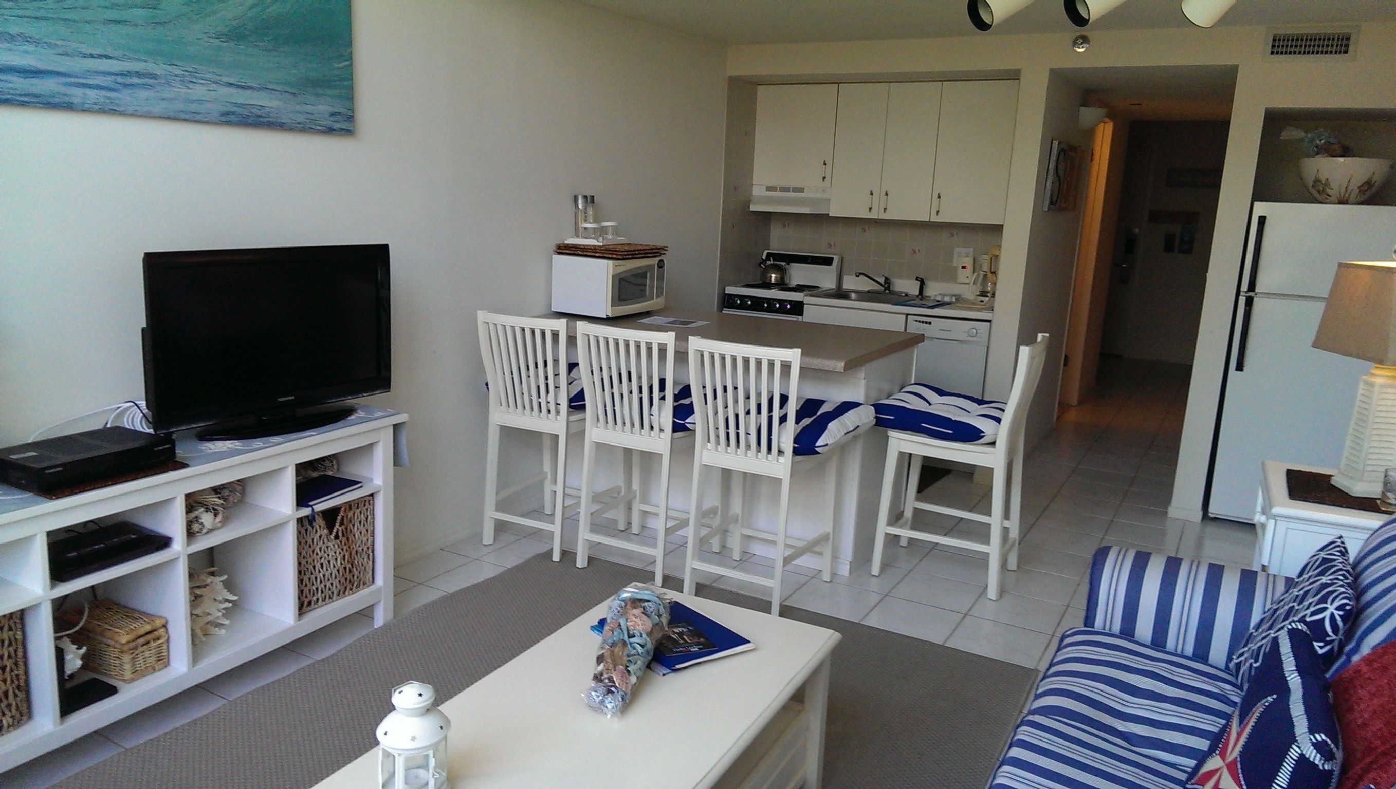 A living room with a TV, white furniture, a kitchen in the background, and a dining area.