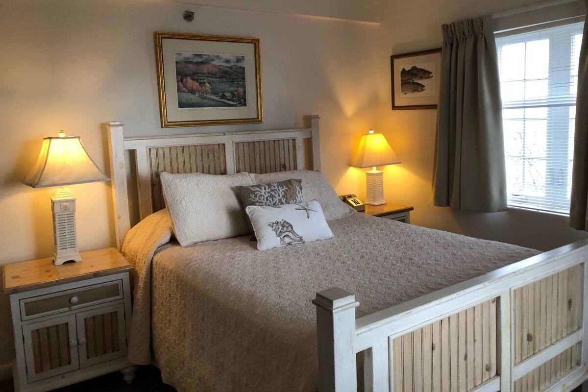 Un dormitorio ordenado con cama, dos mesitas de noche, una lámpara, un cuadro enmarcado en la pared y una ventana con cortinas.