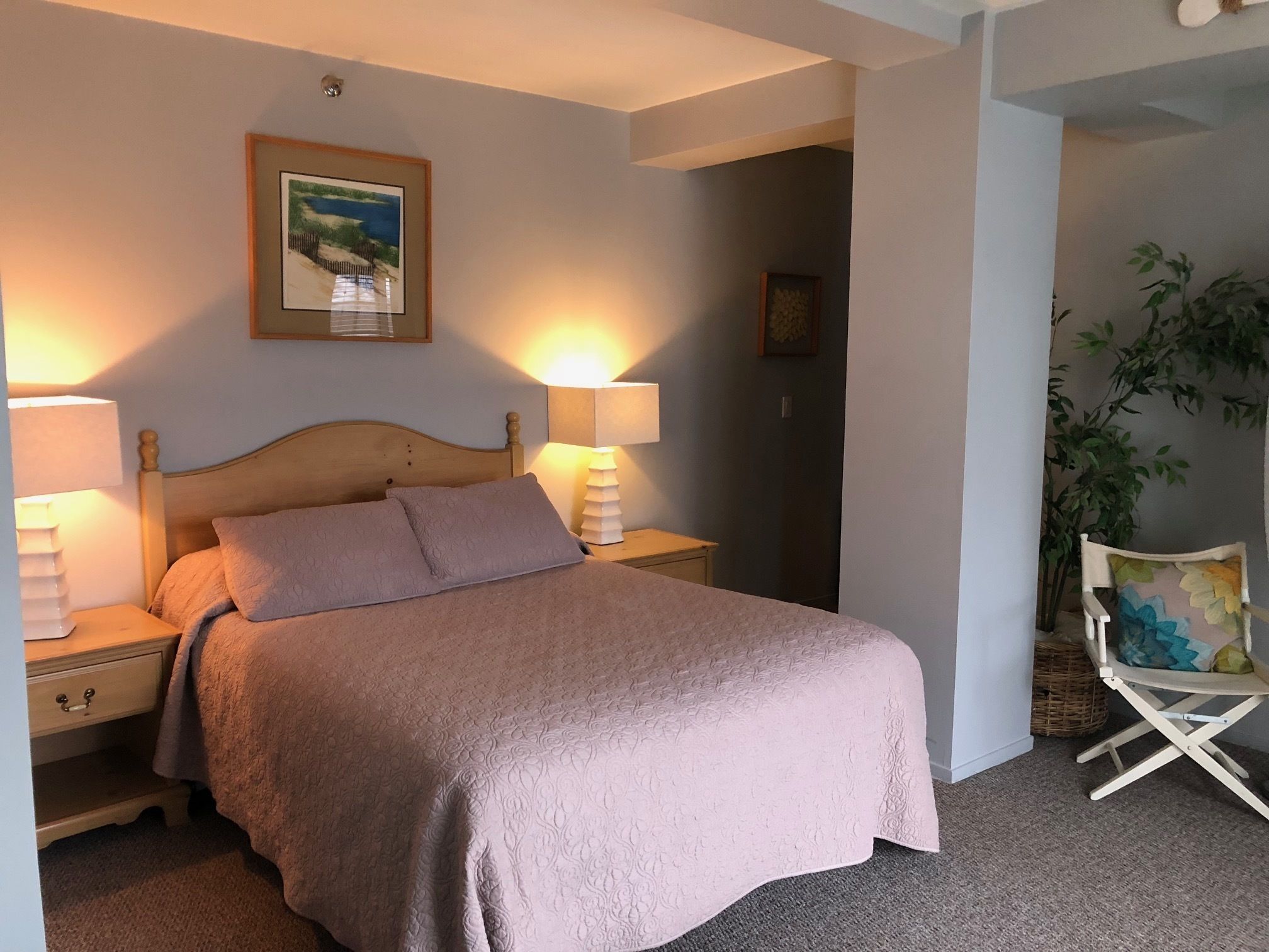 A neatly made bed with a headboard, two bedside lamps, a framed picture on the wall, and a chair with a colorful cushion.