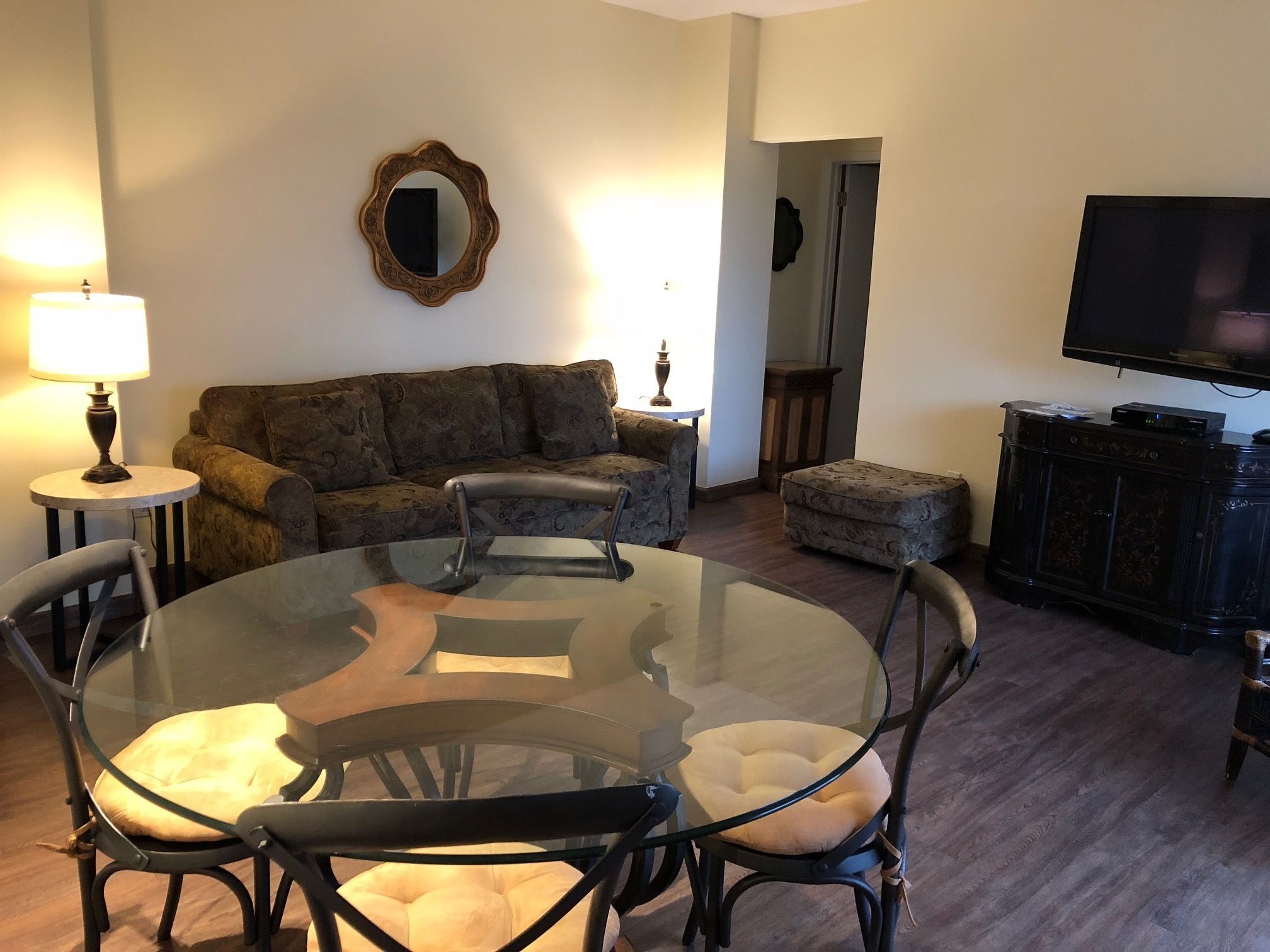 A living room with a glass table, chairs, a sofa, a television, and a lamp.