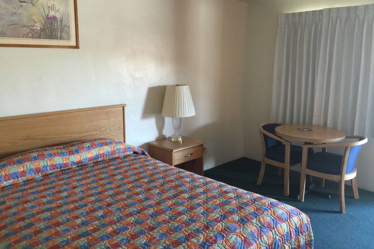 A hotel room with a wooden bed, a colorful quilt, a bedside table with a lamp, a small round table with a chair, and a window with curtains.