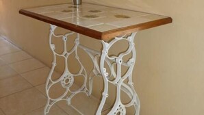 A table with a glass top and ornate white metal legs, placed against a plain wall with a potted plant on top.