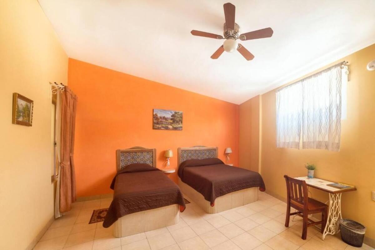 A room with two beds, a ceiling fan, a desk with a chair, and a window with curtains.