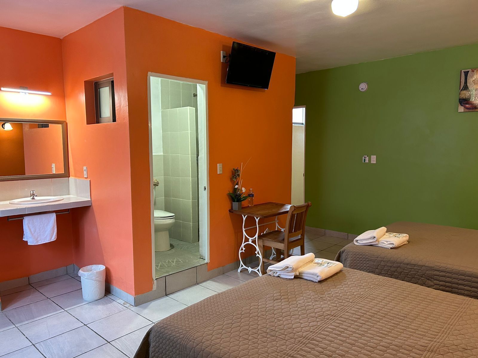 A hotel room with orange and green walls, a bathroom with a white sink and mirror, a desk with a chair, and two beds with towels.