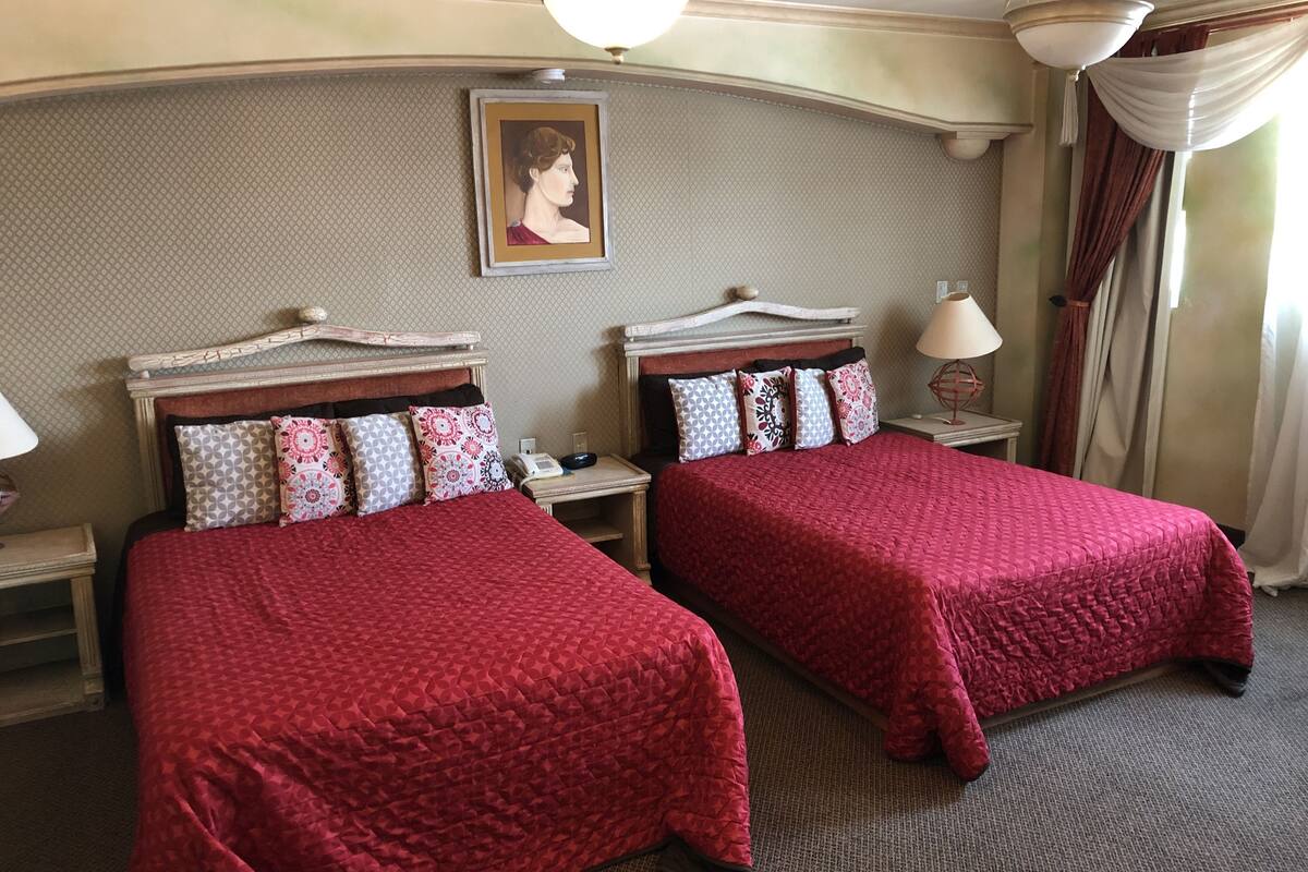 Een hotelkamer met twee bedden, elk met een rode sprei, een nachtkastje met een lamp en een ingelijst schilderij aan de muur.