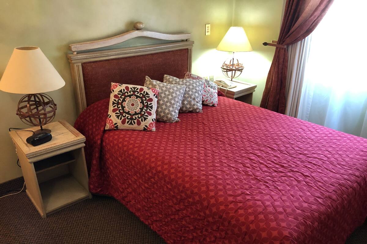 Een hotelkamer met een rood beddeken, meerdere kussens, een houten hoofdbord, bedlampjes en een nachtkastje met een globe lamp.