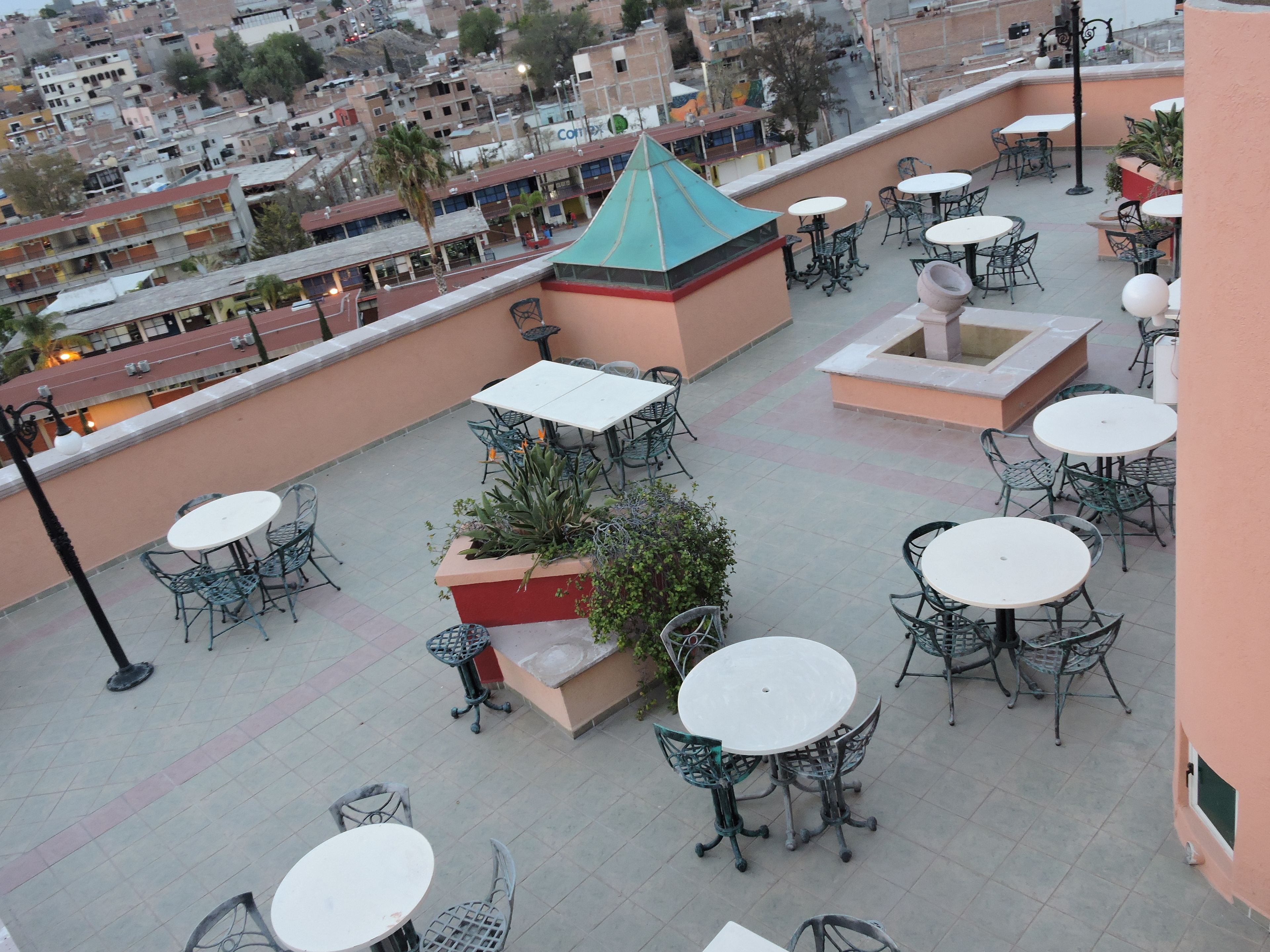 A rooftop terrace with multiple round tables and chairs, surrounded by potted plants and a distinctive green-roofed structure.