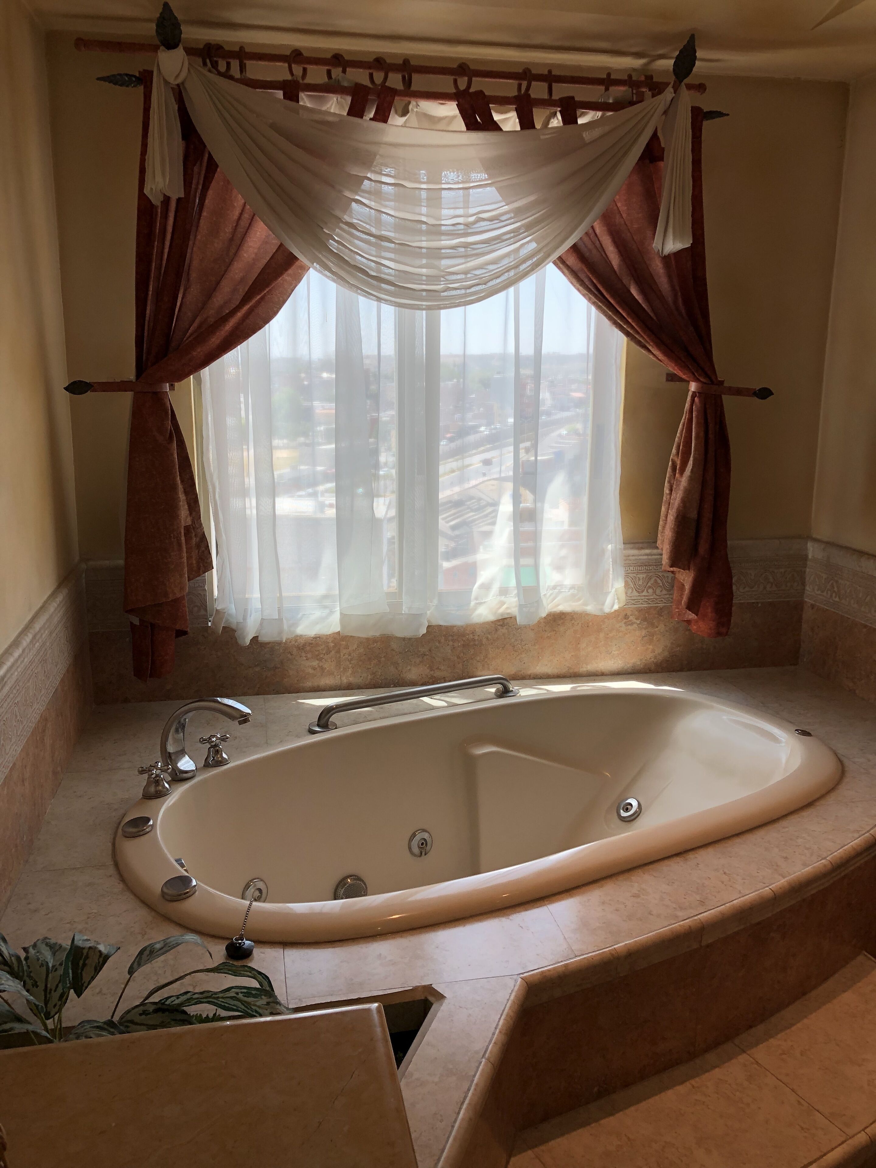 A bathroom with a large bathtub, a window with sheer curtains, and a view of the cityscape.