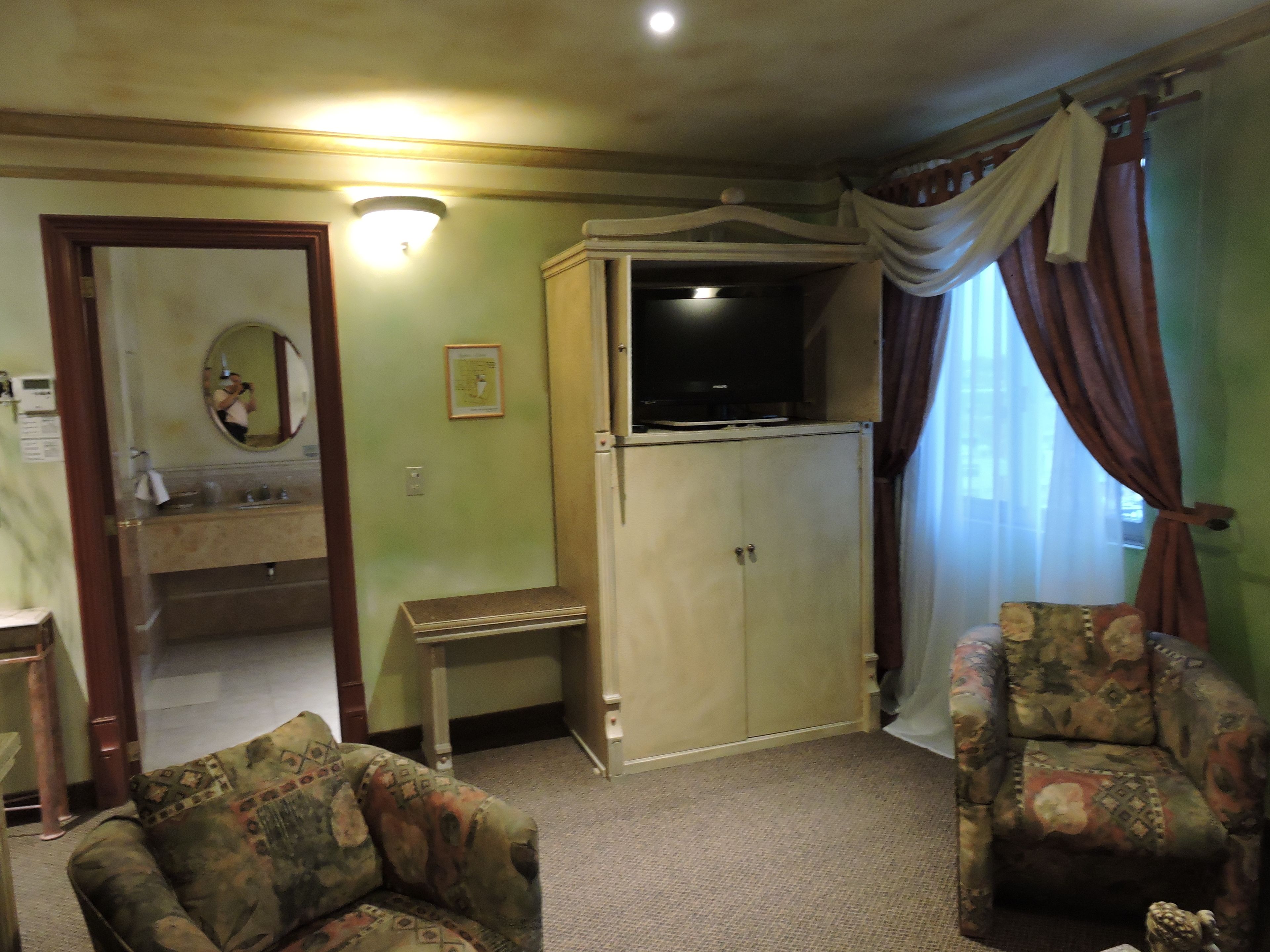 A living room with a television on top of a cabinet, two patterned armchairs, a small table, and a mirror with a picture frame.
